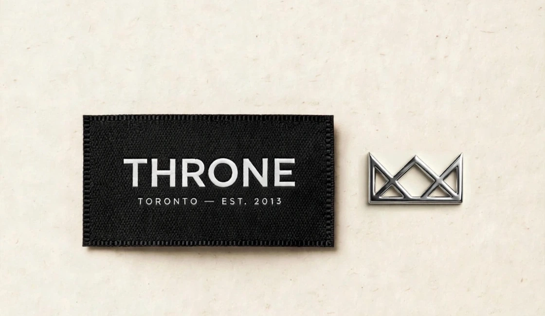 Black woven Throne Toronto label with EST. 2013 beside a small chrome crown pin on cream paper